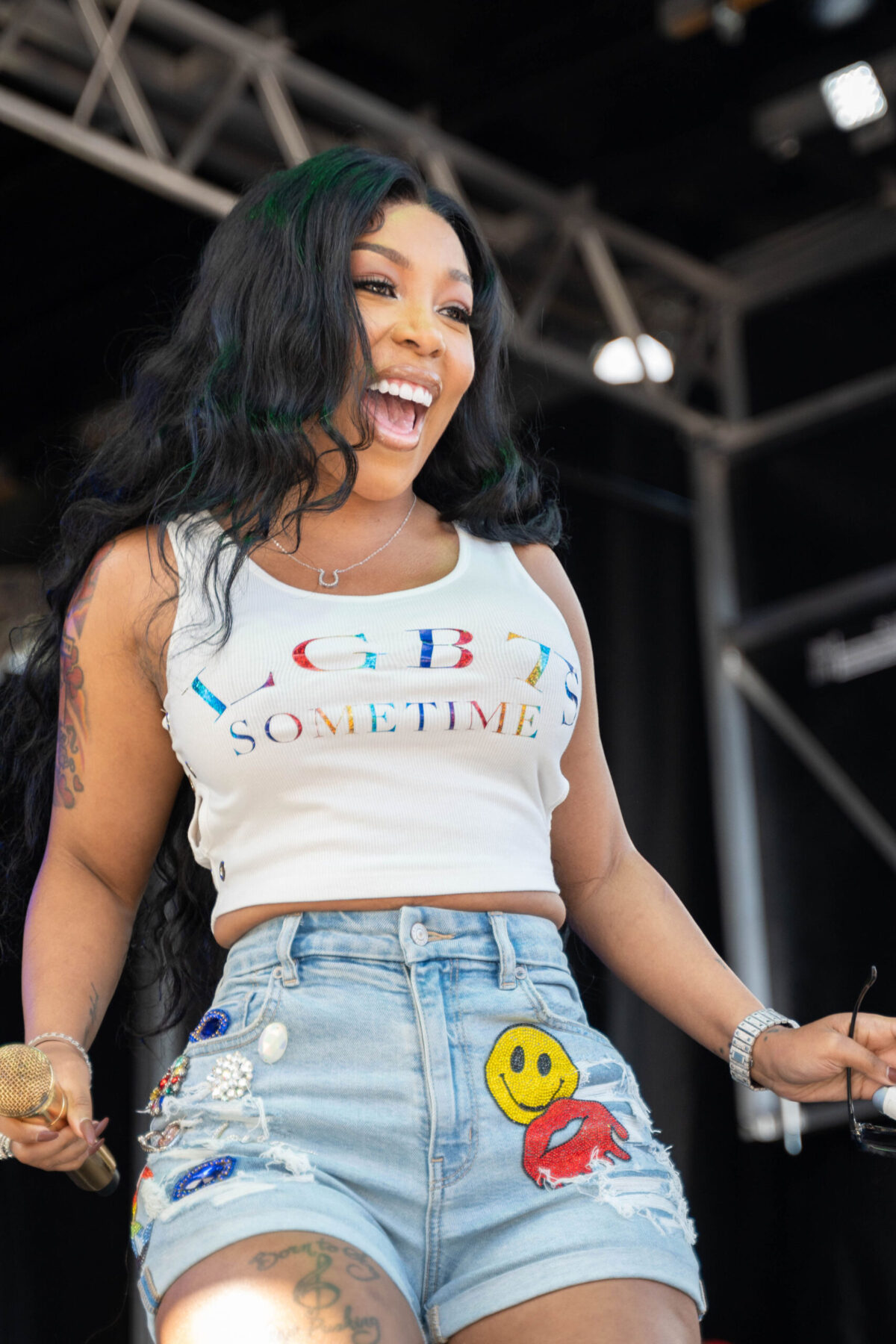 A Black woman stands on stage in a white tank top and jean shorts