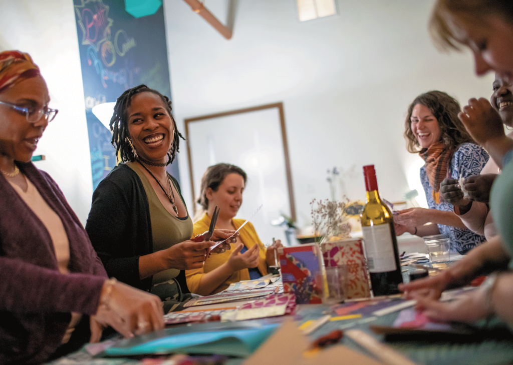 Women smile and laugh around a table, strewn with scraps of paper and cups of wine, while participating in a bookbinding workshop at Drama Mama Bookshop.